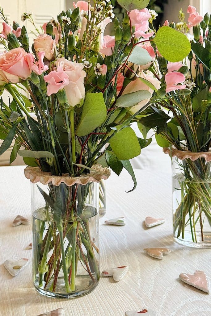 Two glass vases filled with pink roses and greenery are on a white table. Scattered around the vases, along with candles adorned with dried flowers, are small heart-shaped decorations, adding a romantic touch to the arrangement.