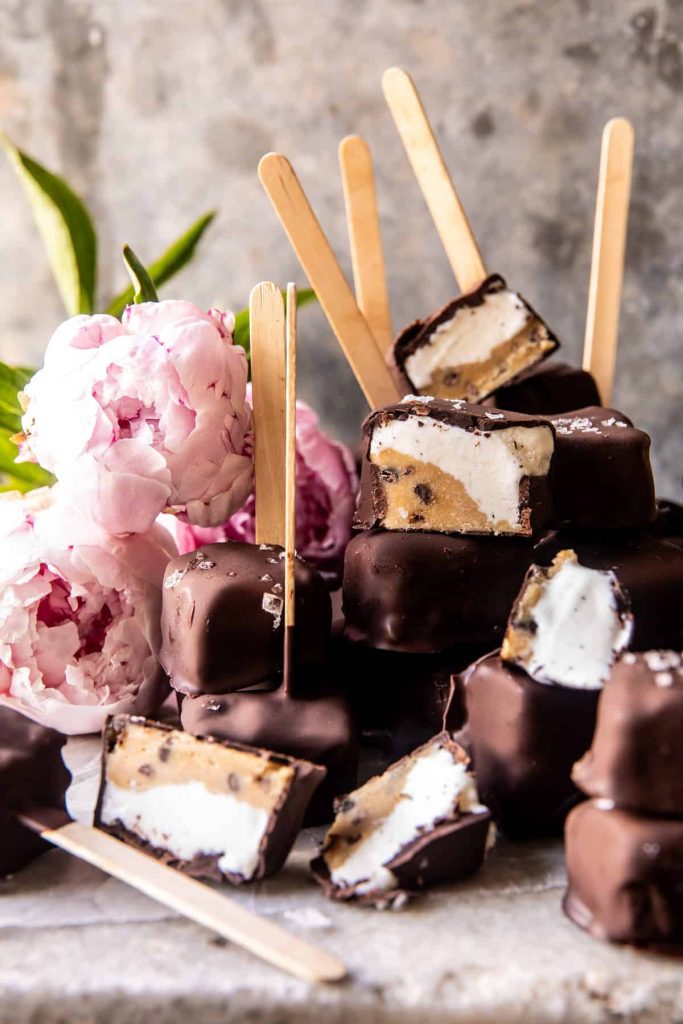 Chocolate-covered ice cream bars, my favorite desserts, reveal enticing layers of vanilla and cookie dough. They are stacked with some sporting wooden sticks. Pink peonies add a splash of color to the rustic setting against a textured surface.