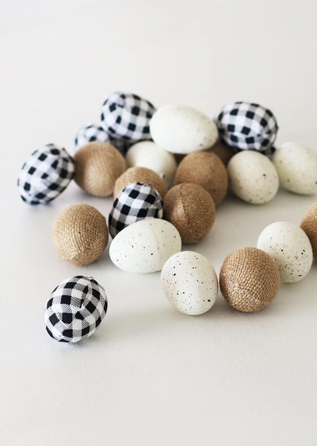 A collection of decorative Easter eggs, embodying my favorite spring crafts, features some wrapped in black and white checkered fabric, others in rustic burlap, and a few painted white with black speckles, all artistically scattered on a white surface.