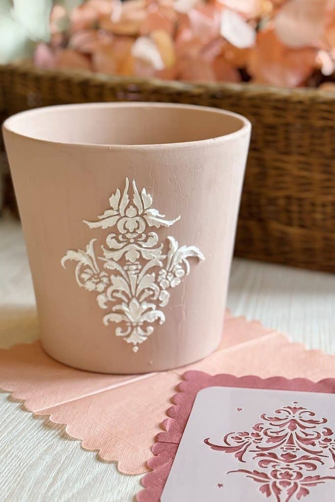 A pink decorative flower pot with a white ornate pattern sits on a light wooden surface, perfect for any DIY enthusiast. It is placed on a pink scalloped mat. In the background, a woven basket holds pink and peach-colored flower petals, much like those found in a lovely garland.