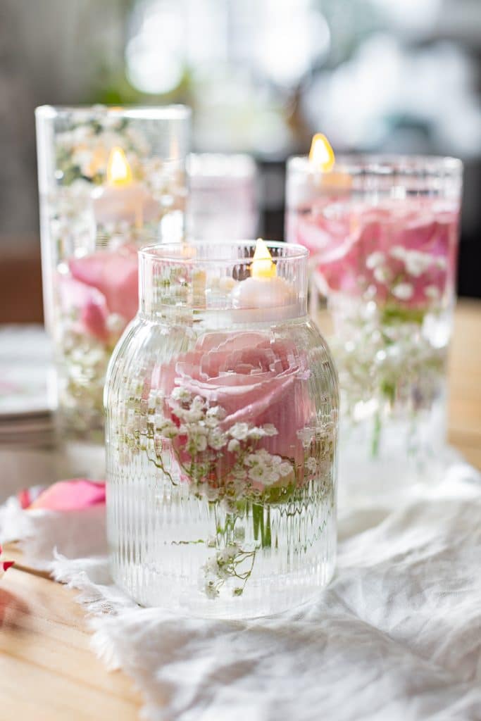 Tall glass jars with lit candles float on water, surrounded by a garland of pink roses and white baby's breath flowers. They rest on a light fabric, creating an elegant and serene DIY centerpiece.