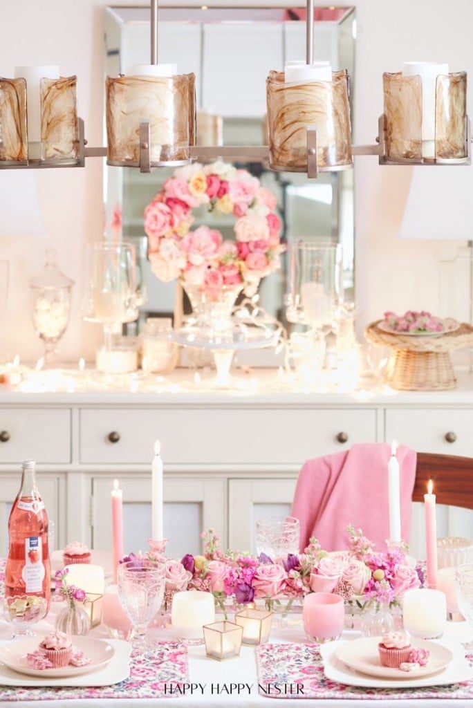 A beautifully set dining table, perfect for Valentine's Table Centerpieces, features pink and white flowers, candles, and a floral tablecloth. A bottle of pink drink adds a touch of romance. A wreath adorned with pink roses hangs in the background, with soft lighting creating a warm, inviting atmosphere.