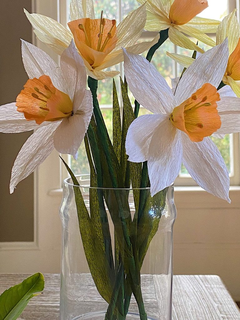 Artificial white and orange daffodils, reminiscent of easy flower paper crafts, sit elegantly in a clear glass vase on a table with a blurred window background.