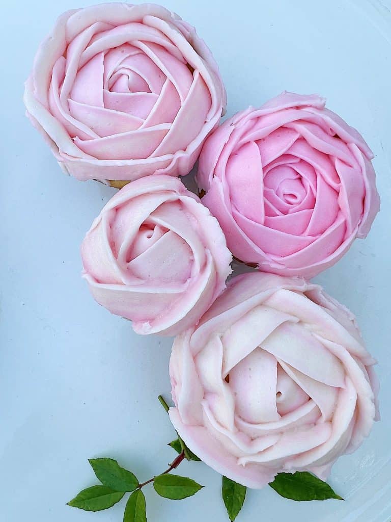 Four cupcakes, my favorite desserts, are topped with intricately piped pink rose-shaped frosting on a white background. A small sprig of green leaves is placed beneath them, adding a touch of natural color contrast.