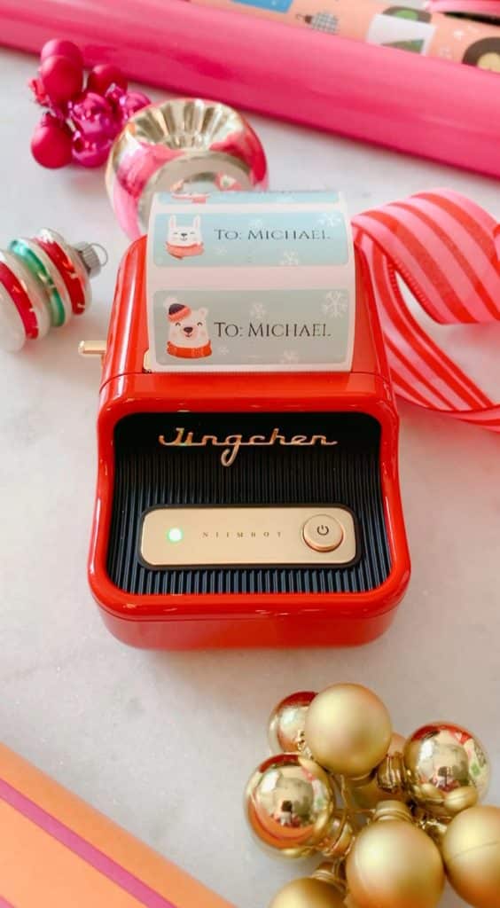 A festive label printer sits amidst a holiday scene, printing "To: Michael" labels with a Santa design. Surrounded by ornaments and shimmering ribbons, it adds a touch of seasonal magic akin to my favorite spring crafts, capturing the joy of creative celebration.