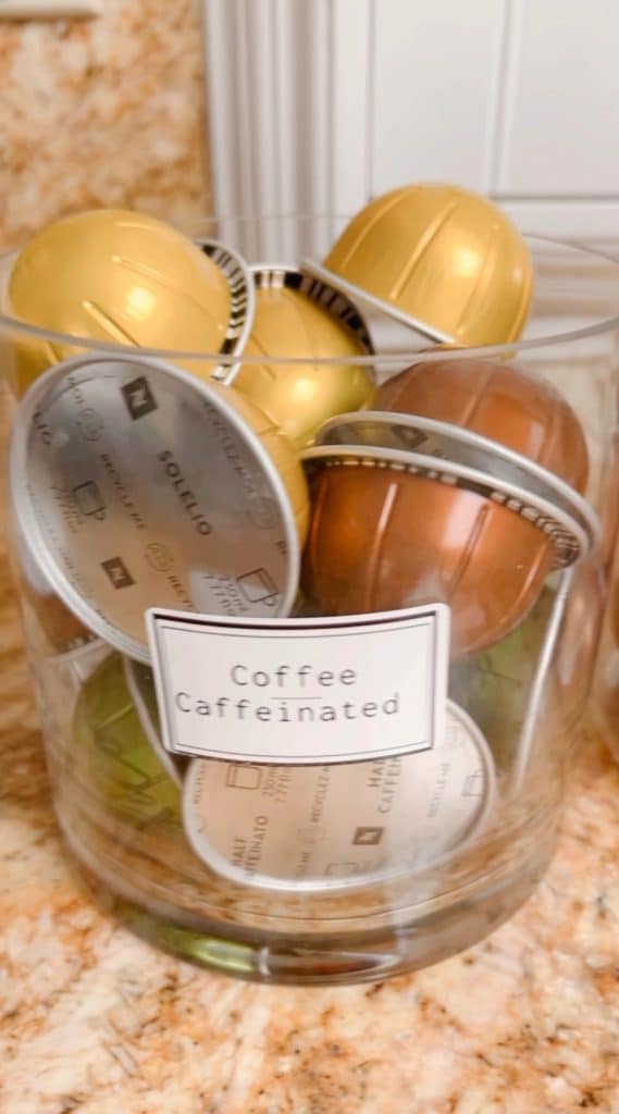 A clear glass container filled with various colored coffee pods, including gold, silver, and bronze, labeled "Coffee Caffeinated," sits on a marbled countertop—a touch of vibrancy similar to my favorite spring crafts.