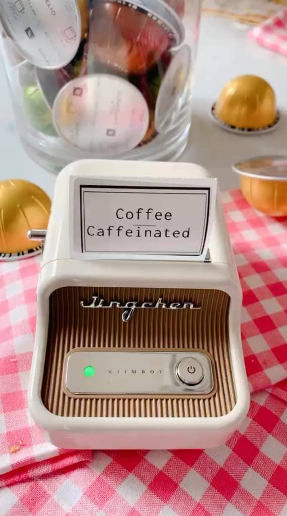 A label printer displays "Coffee Caffeinated" on a pink checkered surface. Behind it, various coffee pods surround a glass jar filled with assorted options, making it feel like one of my favorite spring crafts come to life.