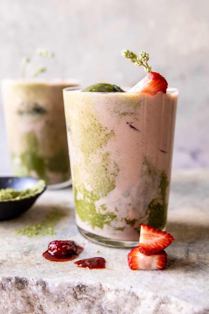 A layered matcha iced latte in a clear glass, adorned with a sprig and a strawberry slice on the rim, exemplifies one of the best summer drinks. Another glass sits in the background, with fresh strawberry slices and a smear of jam on the marble surface beside it.