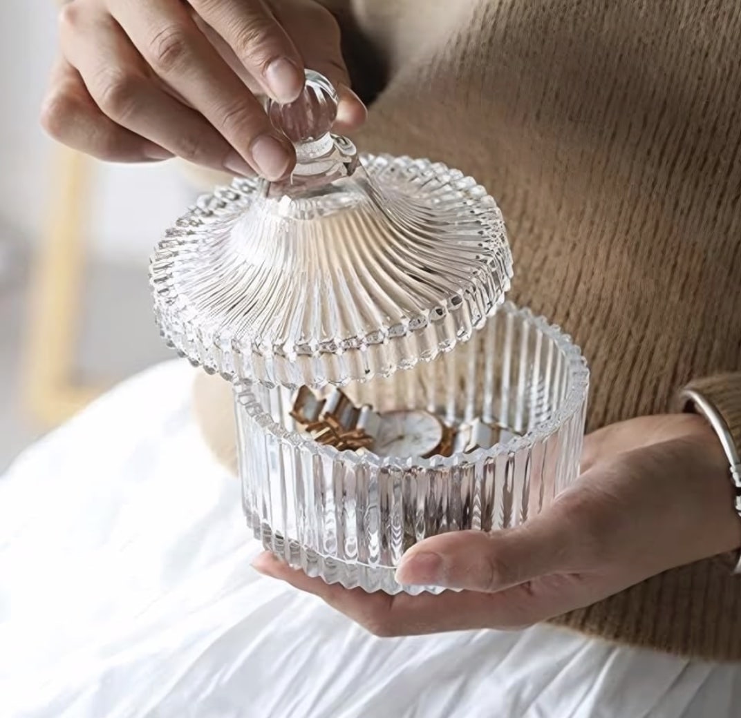 A person holds a round, ribbed glass container with a lid, revealing some gold jewelry inside—one of my favorite spring crafts. They are wearing a beige sweater and a white skirt.