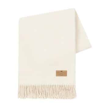 A cream-colored woolen blanket with fringed ends is neatly folded. It has a leather patch label sewn onto one corner.