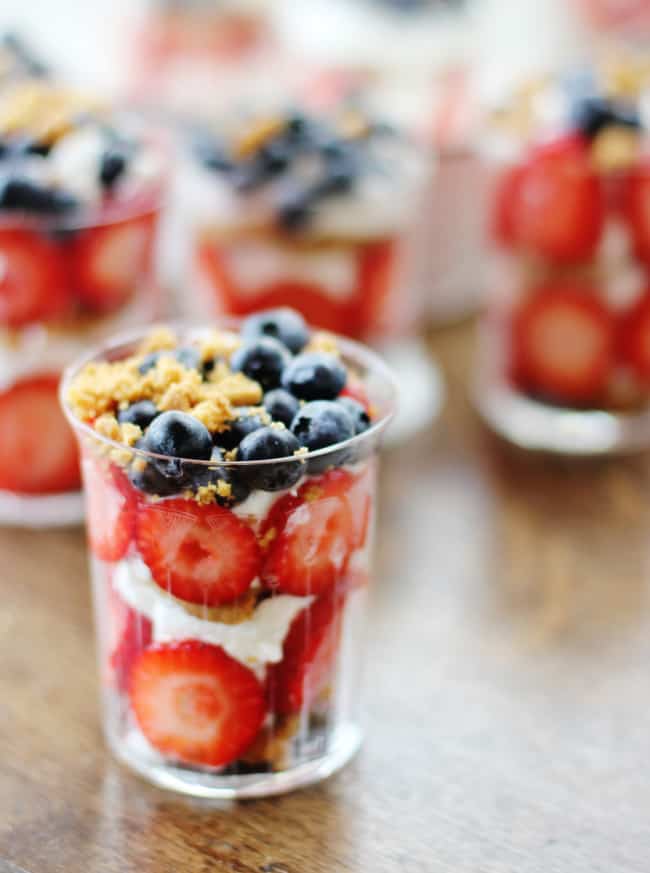 A clear plastic cup filled with layers of fresh strawberries, blueberries, whipped cream, and granola—truly one of my favorite desserts. The vibrant red of the strawberries contrasts beautifully with the deep blue of the blueberries. Other similar cups are elegantly blurred in the background.