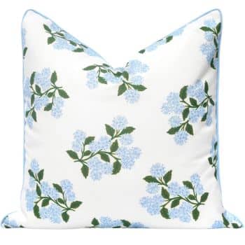 A white decorative pillow featuring a pattern of light blue hydrangea flowers with green leaves. The pillow has a light blue piping along the edges, adding a subtle accent.