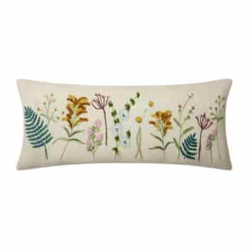 A rectangular pillow with an off-white background featuring an embroidered design of various colorful wildflowers and leaves, including ferns, in green, yellow, pink, and white.