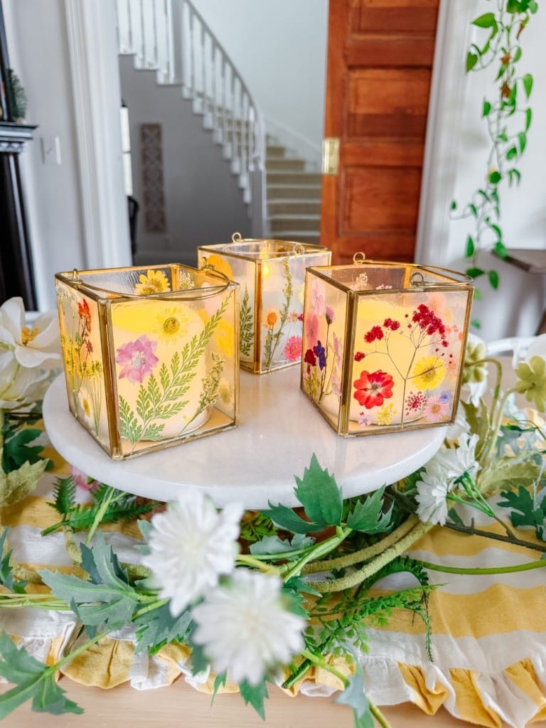 Three square glass candleholders, adorned with floral prints, grace a round white stand. They are nestled amid green leaves and white flowers, creating a DIY garland effect. In the background, a staircase and wooden door add to the picturesque scene.