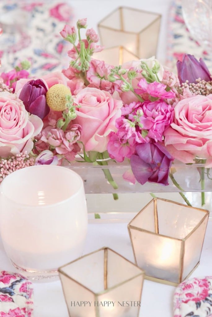 A delicate floral arrangement featuring pink roses, purple tulips, and other pink flowers in a clear rectangular vase, perfect as a Valentine's table centerpiece. Surrounded by decorative candles in gold-trimmed holders on a floral-patterned tablecloth.