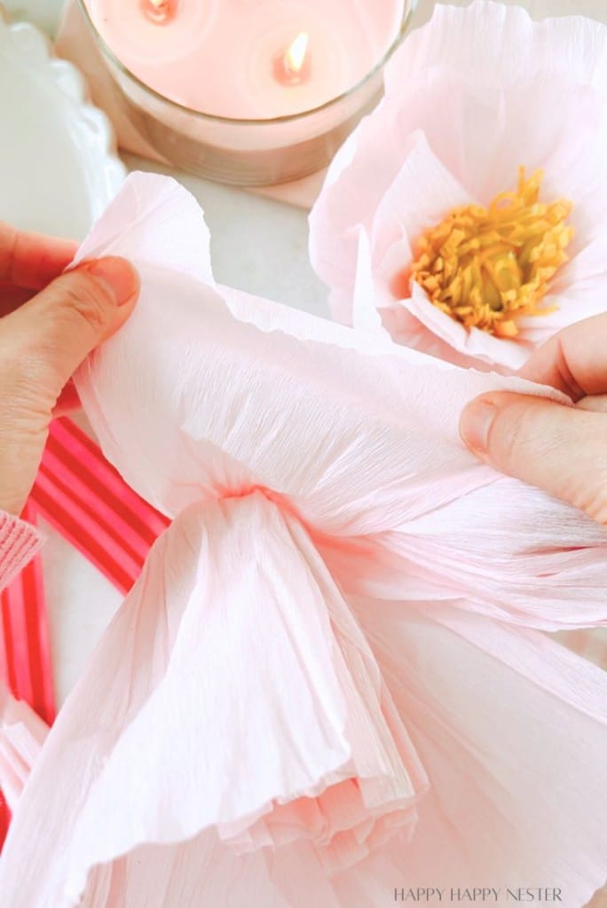 Close-up of hands skillfully crafting a large pink paper flower while a lit candle flickers nearby. A completed bloom and bright pink craft sticks hint at the creation of an enchanting DIY flower garland in the background.