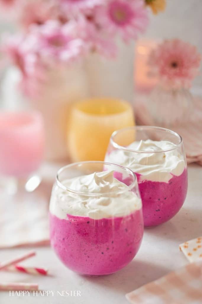 Two glasses of a vibrant pink smoothie, hailed as one of the best summer drinks, are topped with whipped cream and placed on a table. The soft-focus pink flowers and pastel-colored candles in the background create a cozy and inviting atmosphere.