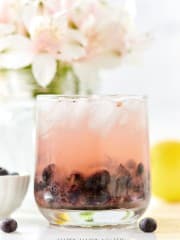 A refreshing glass of iced pink lemonade with blueberries sits on a table, perfect for easy summer mocktail recipes. In the blurred background, a bouquet of white flowers decorates alongside a bowl of blueberries and a bright yellow lemon.