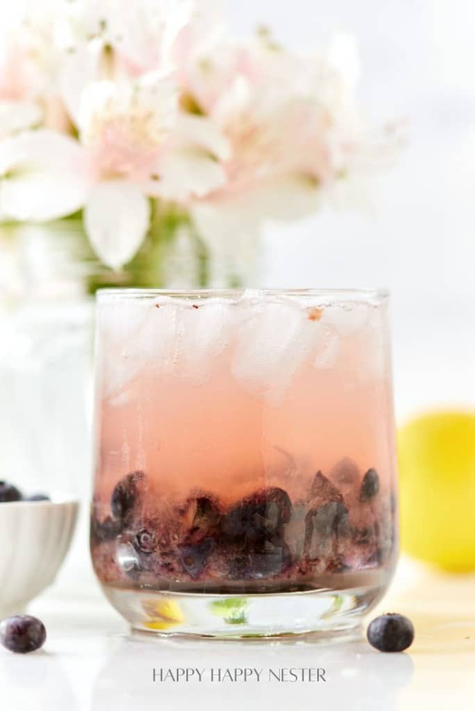 A refreshing glass of iced pink lemonade with blueberries sits on a table, perfect for easy summer mocktail recipes. In the blurred background, a bouquet of white flowers decorates alongside a bowl of blueberries and a bright yellow lemon.