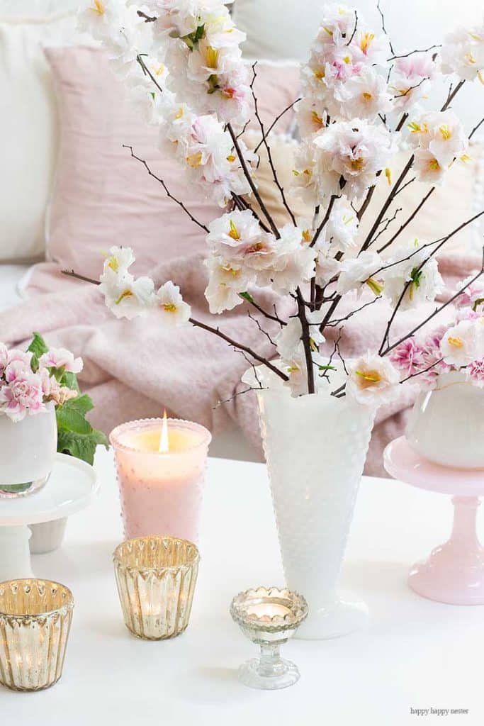 A white vase with cherry blossom branches graces the table, accompanied by a lit pink candle in a matching holder and several decorative gold candle holders. In the background, a pink cushion whispers elegance. The ambiance is soft and pastel, inspiring easy flower paper crafts.