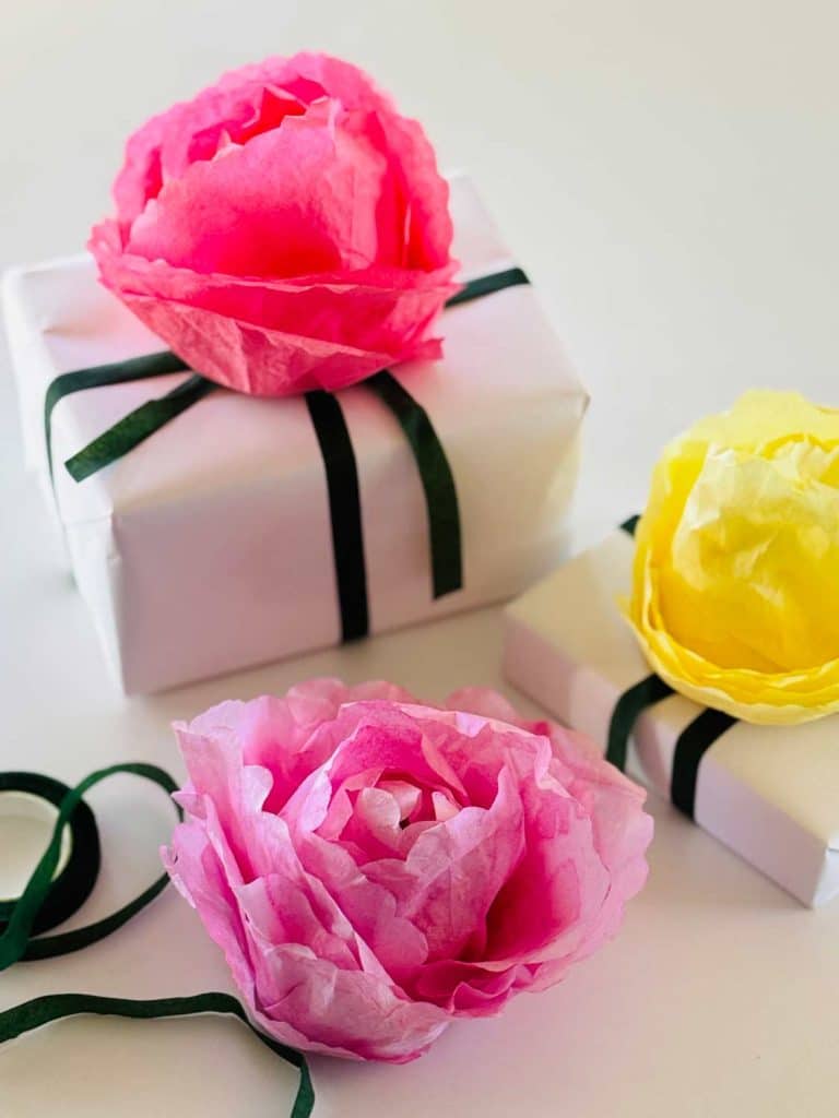 Two wrapped gifts adorned with pink and yellow tissue paper flowers sit elegantly, decorated with dark green ribbons. Nearby, a lovely DIY flower adds a touch of charm to the white surface, creating a beautiful garland effect.