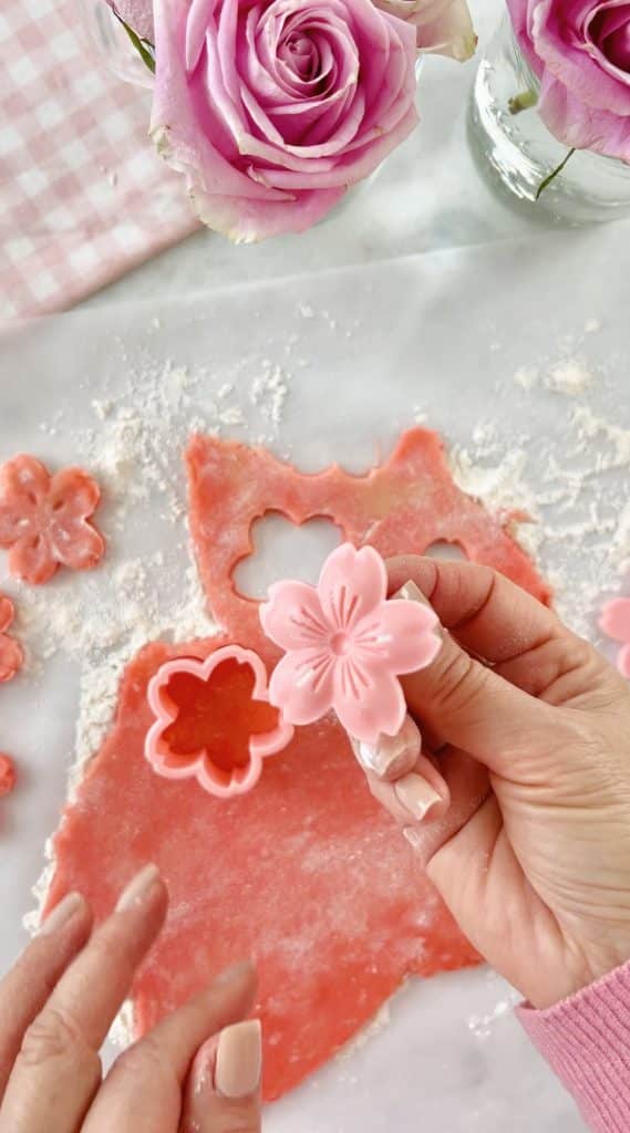 Hands holding a pink flower-shaped fondant cutout over a table with pink dough and flower cutters, crafting one of my favorite desserts. Nearby, pink roses bloom in a vase alongside a charming pink gingham cloth.