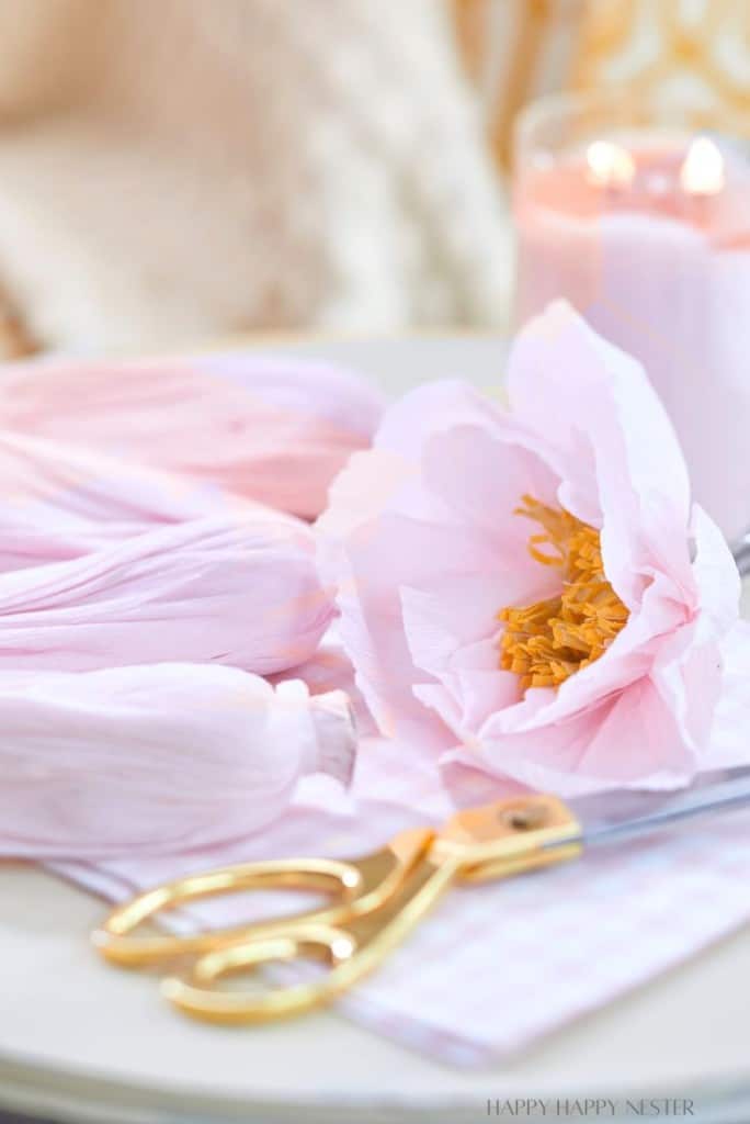 A delicate pink paper flower lies on a table with gold scissors beside it. The background is softly blurred, featuring a pink candle in a glass holder, creating a cozy and artistic atmosphere.