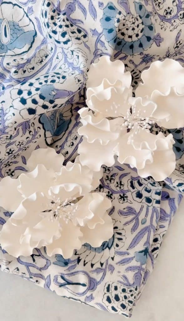 Three white, ruffled fabric flowers, reminiscent of my favorite desserts, sit atop a textured, floral-patterned fabric in shades of blue and purple. The flowers have intricate, wavy petals with small clustered centers, creating a delicate and artistic contrast with the background.