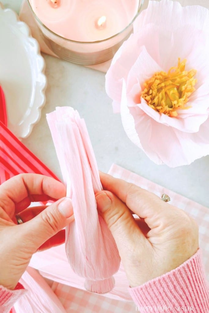 Hands carefully folding pink crepe paper next to a large artificial pink flower, a lit candle, and red striped napkins on a table draped with a pink and white checkered cloth—perfect elements for crafting a DIY flower garland.