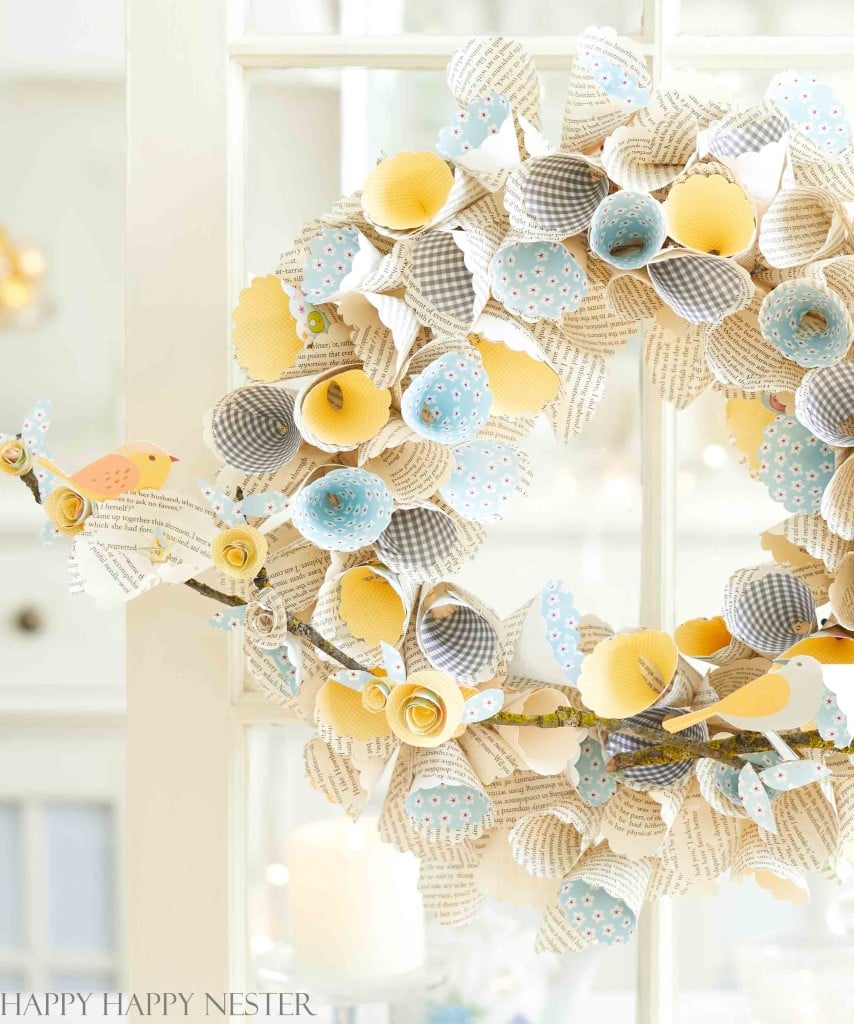 A decorative wreath made of rolled pages and multicolored paper flowers, one of my favorite spring crafts, adorns a glass door. The flowers bloom in shades of yellow, blue, and white with playful patterns. Tiny paper birds perch along the wreath, adding an enchanting touch.