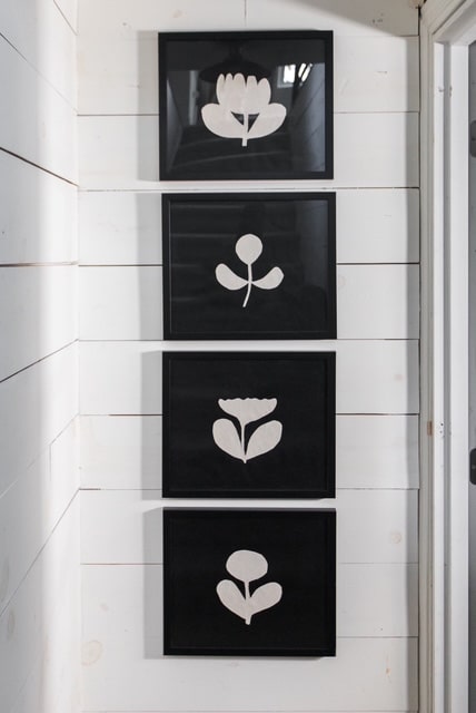 Four framed black and white minimalist floral artworks, reminiscent of a flower garland's elegance, are vertically aligned on a white shiplap wall. Each frame features a distinct simple flower design in white against a black background.