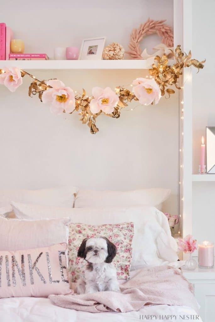 A cozy bedroom features a small dog sitting on a bed adorned with pink and floral pillows. Above, DIY shelves showcase decor items and a handmade garland of pink flowers and golden leaves. A lit candle and soft lighting create a warm ambiance.