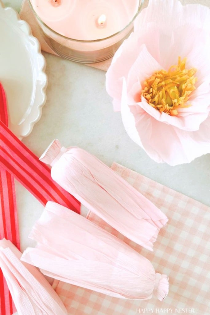 A flat lay image showcases DIY flower garland elements: pink paper flowers, a pink and white checkered cloth, striped pink ribbons, and a white dish. A candle with two wicks peeks in at the top alongside a partially unwrapped paper candy.