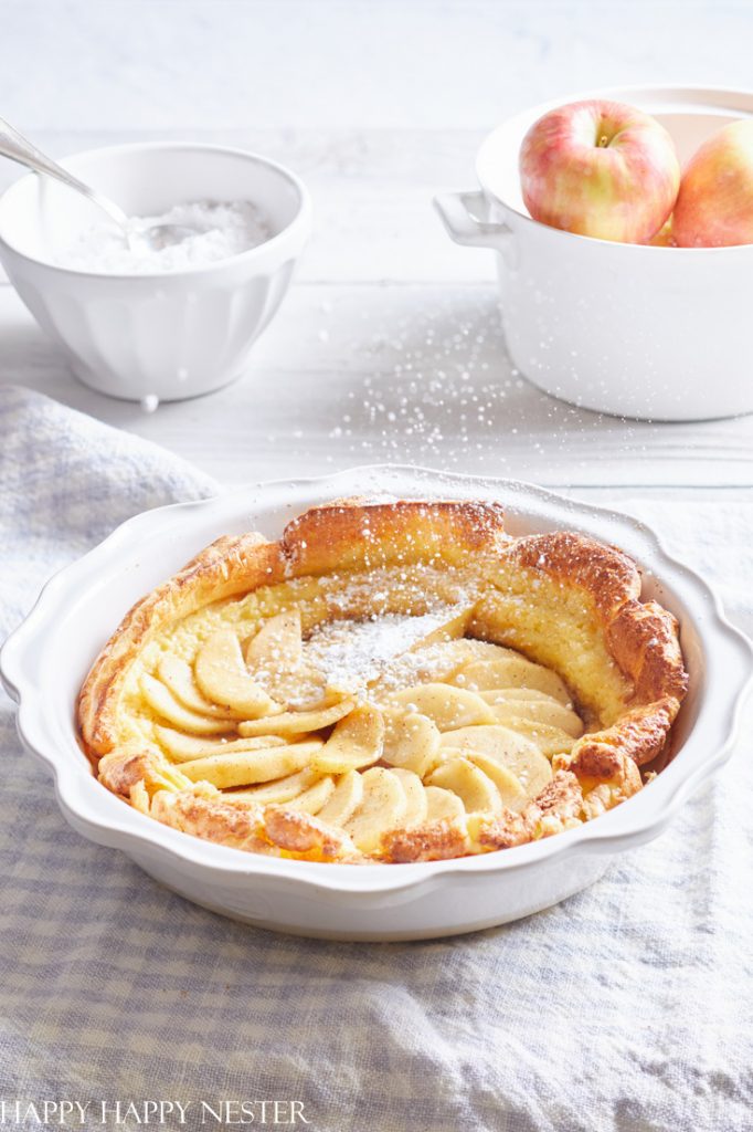 A golden-brown apple pancake in a white pie dish, dusted with powdered sugar, sits among my favorite desserts. Beside it, a white bowl of sugar with a spoon and an apple-filled pot rest on a light wooden surface and checkered cloth.