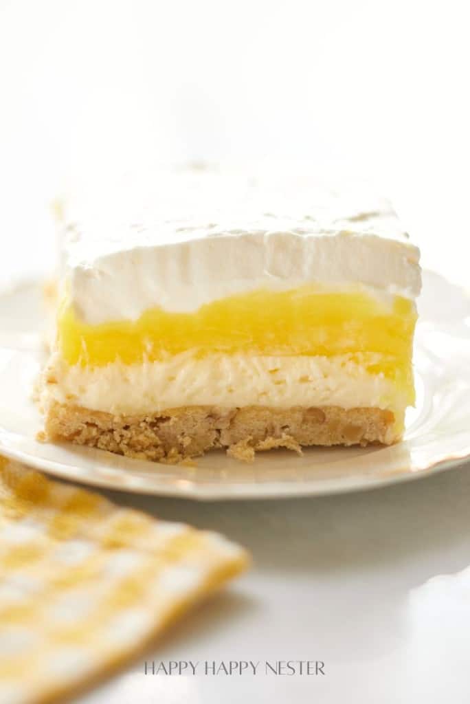 A close-up of a Lemon Lush Dessert slice on a white plate, showcasing its layers of whipped cream, lemon filling, and graham cracker crust. A patterned yellow and white cloth is partially visible in the foreground, adding a touch of charm to this delightful recipe.