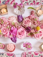 A Valentine’s table centerpiece featuring pink roses, purple tulips, and various pink blossoms, interspersed with green leaves. Surrounded by lit candles in geometric holders, pink cupcakes, and a floral-patterned tablecloth, it creates a romantic ambiance perfect for the occasion.