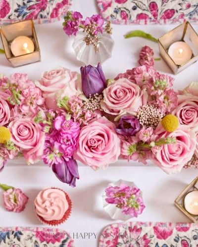 A Valentine’s table centerpiece featuring pink roses, purple tulips, and various pink blossoms, interspersed with green leaves. Surrounded by lit candles in geometric holders, pink cupcakes, and a floral-patterned tablecloth, it creates a romantic ambiance perfect for the occasion.