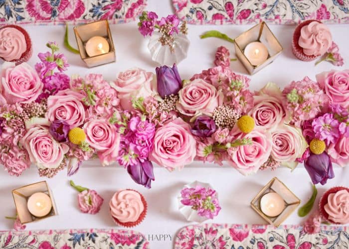 A Valentine’s table centerpiece featuring pink roses, purple tulips, and various pink blossoms, interspersed with green leaves. Surrounded by lit candles in geometric holders, pink cupcakes, and a floral-patterned tablecloth, it creates a romantic ambiance perfect for the occasion.