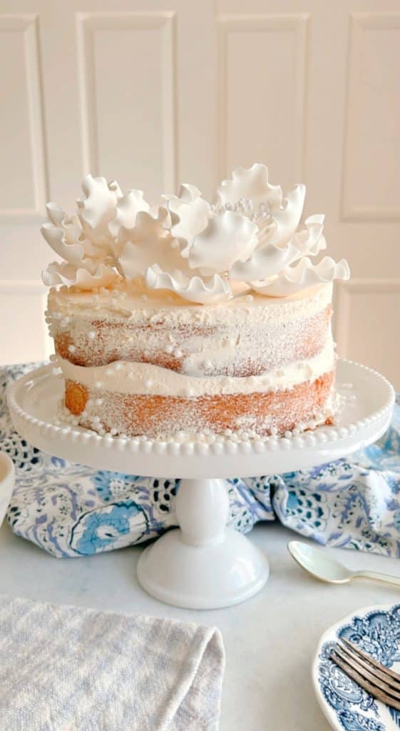 A semi-naked cake on a white pedestal stand, adorned with delicate white icing flowers on top, is truly one of my favorite desserts. The cake is set against a patterned blue and white tablecloth, creating an elegant presentation.