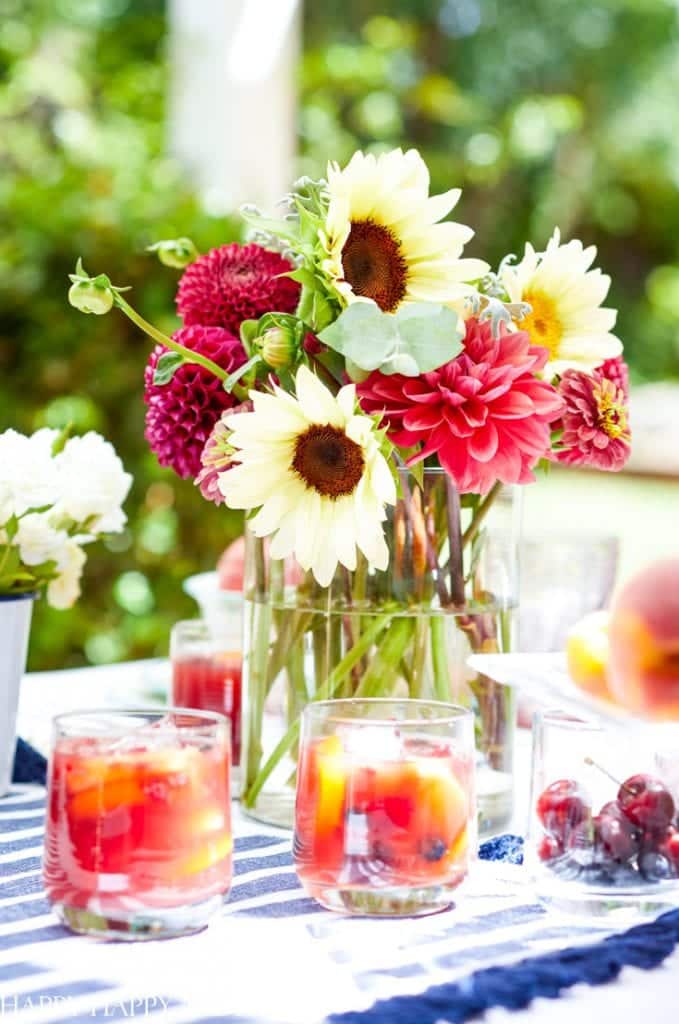 A colorful bouquet of sunflowers and red dahlias in a glass vase sits on a table. In the foreground are glasses of the best summer drinks, infused with fresh cherries and peaches. A blue and white striped cloth adds a summery vibe.