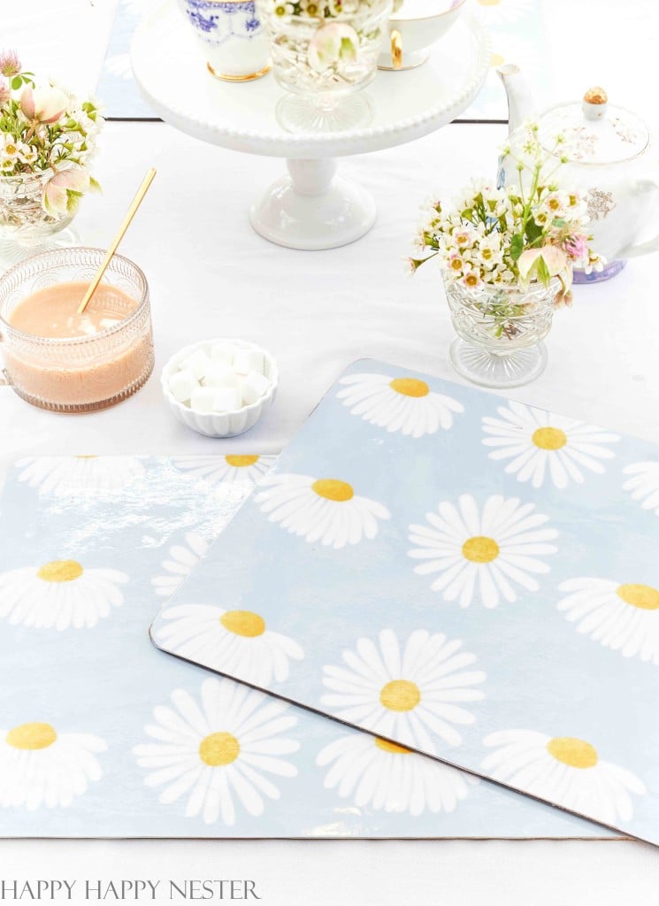 A table set for tea with a daisy-patterned placemat, floral teapot, cups, and a glass of peach-colored drink with a gold spoon showcases my favorite spring crafts. Small vases of white flowers and a bowl of sugar cubes complete the setting.