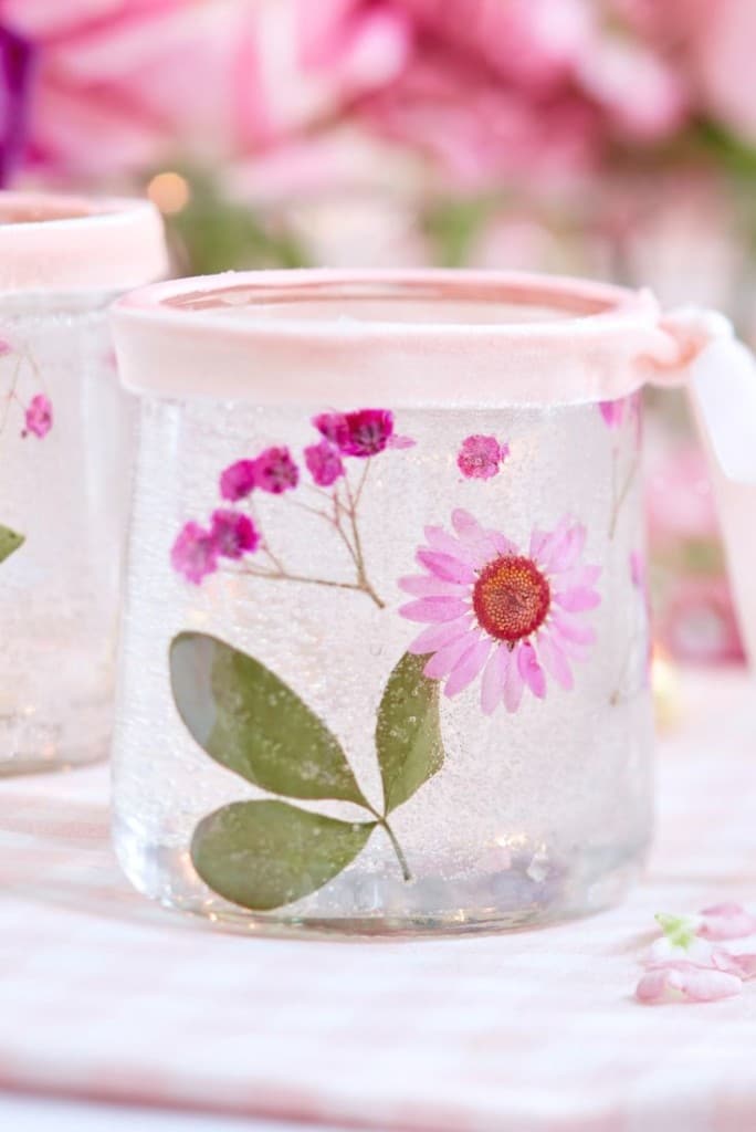 A glass jar, one of my favorite spring crafts, is adorned with pressed pink flowers and green leaves, featuring a soft pink rim. The background is softly blurred with more flowers and a checkered tablecloth.