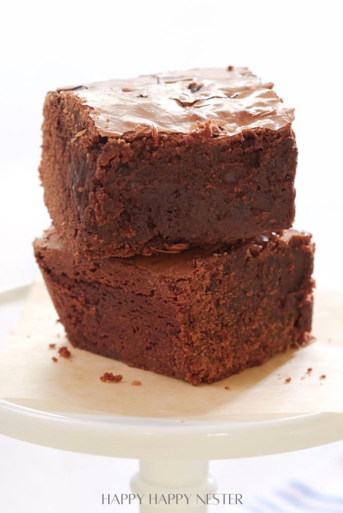 Two thick, fudgy chocolate brownies stacked on a white cake stand, boasting a rich, glossy top layer. These are truly my favorite desserts.