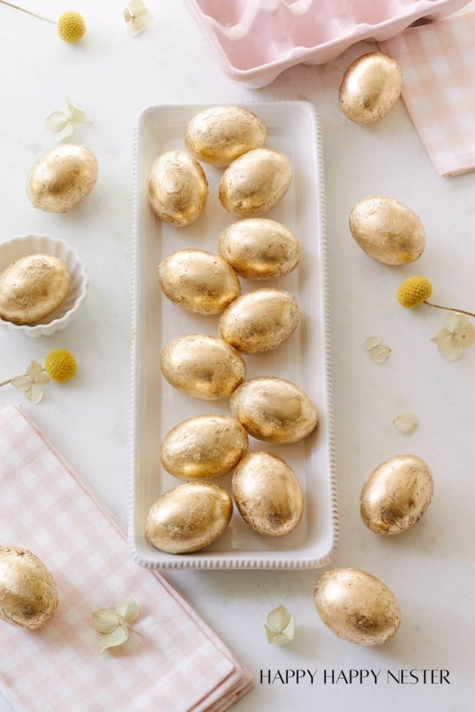 A rectangular white dish holds shiny gold eggs, reminiscent of my favorite spring crafts, arranged neatly. Surrounding the dish are yellow pom-pom flowers and small white blooms, all atop a pink checkered cloth. A pink egg carton peeks out from the background.