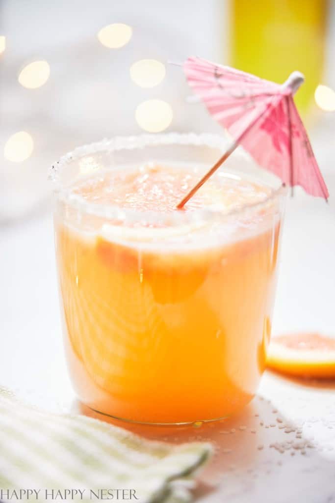 A refreshing orange drink in a clear glass with a sugared rim, garnished with a pink paper umbrella—it captures the essence of best summer drinks. The scene is completed by a light and airy background, with a green striped cloth at the bottom.