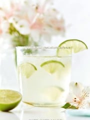 A glass of a lime cocktail with ice and lime wedges on the rim sits on a white surface, embodying the essence of easy summer mocktail recipes. A halved lime and light pink flowers are blurred in the background, setting a refreshing scene.