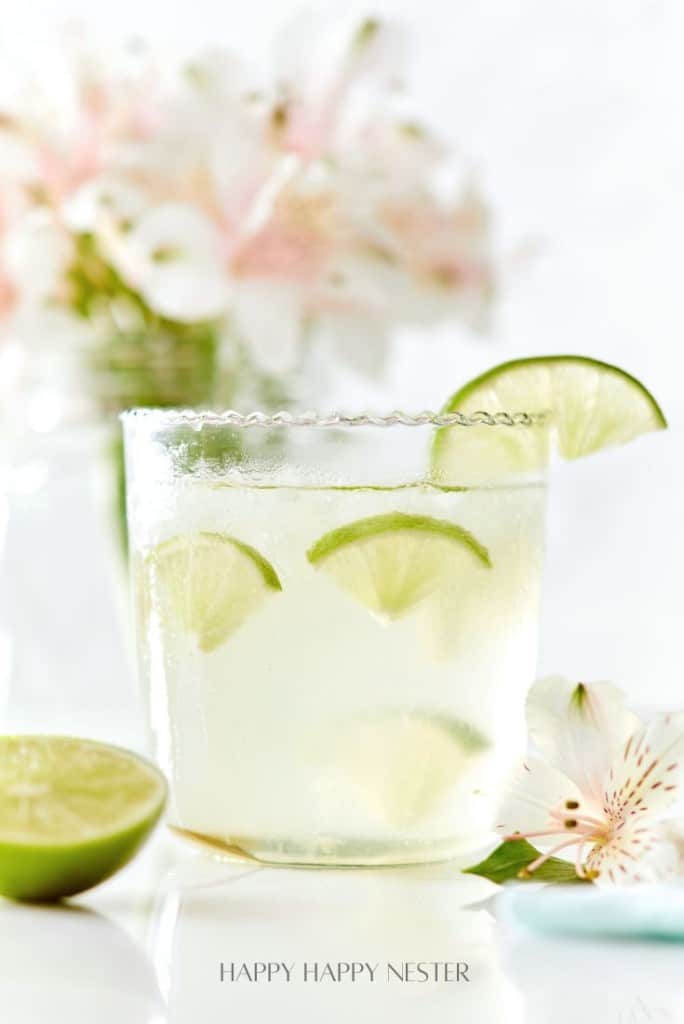 A glass of a lime cocktail with ice and lime wedges on the rim sits on a white surface, embodying the essence of easy summer mocktail recipes. A halved lime and light pink flowers are blurred in the background, setting a refreshing scene.