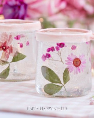 Two decorated glass jars adorned with pressed flowers and leaves, perfect for making candles with dried flowers. Topped with pink fabric and tied with ribbons, the jars sit on a checkered surface. A blurred floral background completes the soft, romantic atmosphere.