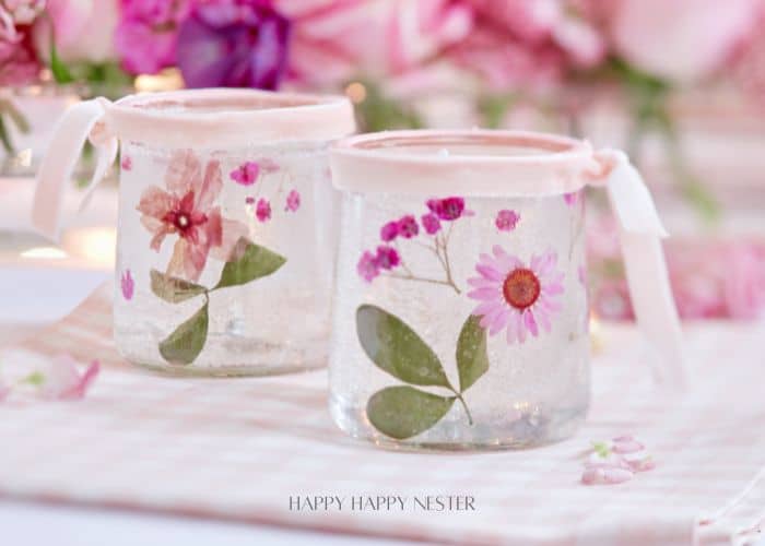 Two decorated glass jars adorned with pressed flowers and leaves, perfect for making candles with dried flowers. Topped with pink fabric and tied with ribbons, the jars sit on a checkered surface. A blurred floral background completes the soft, romantic atmosphere.