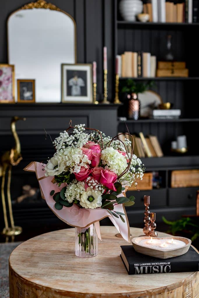 A bouquet of pink and white flowers in a vase sits on a wooden table adorned with candles surrounded by dried flowers and books. The background features a dark bookshelf with various books and decorative items. A mirror and small framed pictures are visible on the wall.